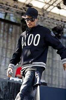 T.I. walking onto a stage wearing a cap, hoodie, jeans and sunglasses