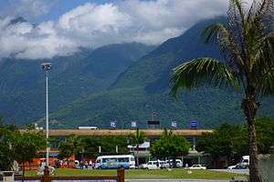 The front of Hualien station with a palm tree in the foreground and a big mountain with clouds in the background behind the station.