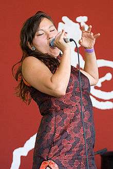 "A colour photo of Tagaq on stage singing. She is holding a microphone while wearing a red and black dress."