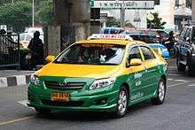 A Toyota saloon/sedan driving on the road, its top half painted in yellow and the bottom half in green, with a sign on the roof saying "TAXI-METER" and a yellow licence plate