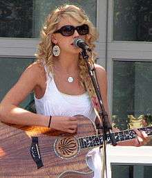 Taylor Swift, wearing a white dress and sunglasses, plays an acoustic guitar while standing at a microphone stand