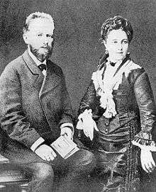 A middle aged man with dark hair and a beard, wearing a dark suit and holding a book, sits next to a young woman in a black dress wearing her hair up on her head