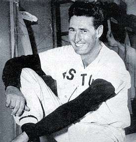 A smiling man in a white baseball uniform with a black long-sleeved shirt underneath sits with his arms around his right leg.