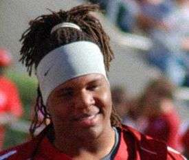 An American football player smiles wearing a red jersey and white headband.