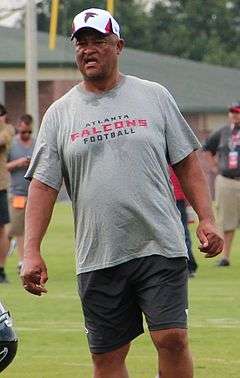 Candid knees-up photography of Robiskie on a football practice field wearing a grey Atlanta Falcon t-shirt, dark grey shorts and a white Falcons baseball cap