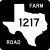 Image of FM 1217 highway shield. The square shield has a white symbol in the shape of Texas as the state appears on maps on a black background. Inside this symbol is the number 1217. The black background contains the words FARM in the upper right corner and ROAD in the lower left corner, both in white capital lettering.