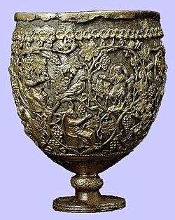 A photo of a large ovoid vessel standing on a short knobbed stem. The cup comprises a silver body enclosed in an openwork layer of gold. The gold ornamentation represents vine scrolls enclosing small seated and praying figures.