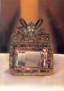 A large rectangular pink multifaceted gemstone, set in a decorative surround. The decoration includes a row of small clear faceted gemstones around the main gem's perimeter, and clusters of gems forming a crest on one side. The crest comprises a three-pointed crown faced by two unidentifiable animals.