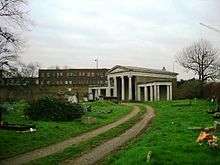 A double track leads through grass towards a small Classical-style building. The central portion has four columns supporting a pediment, and wings with columns extend from each side.