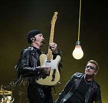 The Edge and Bono clothed in leather jackets, as the Edge holds a guitar vertically. A large dangling light bulb hangs between them.