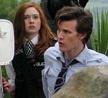 A woman with red hair wearing a policewoman's costume looks alarmed. In front of her, a dark-haired man in a tattered shirt and tie looks to the left.