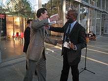 A white man wearing a gray suit reaches to embrace Jones, while holding a book in his right hand. Jones, who is also reaching out, wears a dark suit and has a microphone and piece of paper in his left hand. Inside a glass-walled building behind them, a display says "Climate is an angry beast and we are poking at it with sticks".