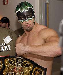 A topless Caucasian man, with green hair and wearing a black and white mask that covers the top half of his face, poses with his arm raised across his chest and his hand in a fist. A wrestling championship with a black strap is visible in the foreground of the image, and is inscribed with "WWE Tag Team Championship".