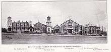 A black and white photograph showing the buildings built/designed by Stanley. From left to right they are the Technical School, the small Hall, the Clock Tower, the large Hall and the Secretary's House. They are seen from across the road, with fencing in front of the buildings.