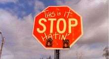In the photo, a red stop sign can be seen. On the stop sing, the word "Stop" is in the center, and written above it are the words "This Is it" and at the bottom of the sign, the word "Hatin'" is written in capital letters; all the words on the sign are written in yellow. In the background, a blue sky, that is mainly full of white clouds, is shown and a metal poll is shown behind the sign.