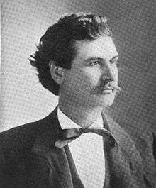 A man with bushy, black hair and a thin mustache wearing a black jacket, vest, and tie and a white shirt