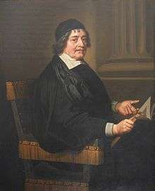 A man with a black cap and gown, seated.  He is holding an open book in his right hand and pointing to it with his left