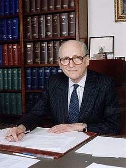 Lord Bingham sitting at his desk with pen in hand and two large bookcases behind him