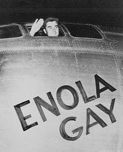 A man waves from the cockpit window of a plane with the words "Enola Gay" written on it