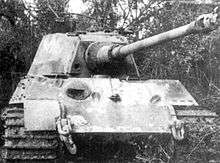 A head-on view of a large tank with a flat-faced turret. Its sloped bow armour is scarred with several fist-sized dents, and there is a fist-sized hole in the front of the turret