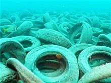 A bed of skummy tires rests piled upon the ocean's floor; a small yellow fish swims by the left.