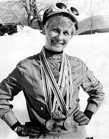 A smiling short-haired blond woman wears a track suit with gloves, and a hat with white skiing goggles. She holds her hands on her waist and carries two medals around her neck. Behind her, a sunny and snowy landscape.