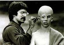Savini, on the left, applies make-up behind Lehman's ear. Lehman's bald head has been made to appear over-large; his eyes point in different directions, and his teeth are extremely crooked.