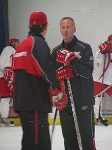 A man wearing a black shirt & black pants is standing on a hockey rink. He has red gloves and is holding a stick. He is talking to a man whose back is to camera.