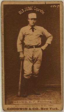 A baseball player is standing, holding a baseball bat down his right side.