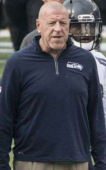 Candid waist-up photograph of Cable wearing a dark blue, long sleeve pullover with a Seattle Seahawks logo standing on a football sideline