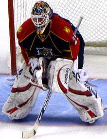 An ice hockey goaltender stands in the crease he is bent over at the waist with his arms on top of his leg pads. The pads are mostly white with stylized red swoops going through them, he wears a matching glove and blacker. His hockey jersey is a navy color with red shoulders and a yellow outline. On his chest is the Panthers' logo of a leaping panther with only his head visible.