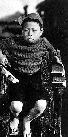 A young Japanese boy of about nine, scowling and staring towards left of frame, wearing a sweater and short pants and sitting on what appears to be a fence, with out-of-focus houses seen in the background.