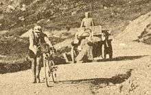 A man walking uphill, alongside his bicycle, followed by a car.