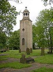 A slender stone tower in a graveyard. On the left face are two windows, one round-headed, the other round; on the right face is a clock; and on the summit is a cupola surmounted by a ball finial