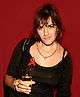 Tracey Emin, holding a wine glass filled with a translucent, peach-colored liquid. She is wearing a black top, with a red ribbon attached.