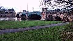 A bridge with three arches spanning a wide river. The near bank is grass with some trees bare of leaves. The sky is blue with many white clouds.