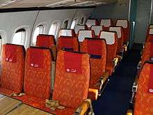 The passenger cabin of an airliner with orange-coloured seats and blue carpeting, the cabin is illuminated by daylight from the windows.