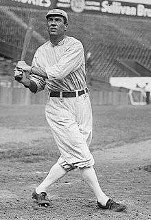 A man wearing an old-style white baseball uniform swinging a baseball bat