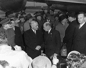 Truman and Attlee in dark suits shake hands. There are surrounded by a crowd of people, all dressed warmly. In the background is a propeller driven airliner.