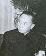 A balding man looking to the left, dressed in a dark jascket buttoned to the neck