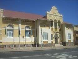 A yellow and white neo-classicist building in bright sunlight. The roof of corrugated iron sheets is painted dark red, on the main gable the inscriptions read "A.D.1913" and "SADC Tribunal". Seven flagpoles with national flags of countries from the Southern African Development Community are mounted onto the pavement in front of the building.