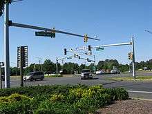 Several cars pass through a signalized intersection viewed from behind planted shrubs.  Small green signs on the traffic light pole masts indicate the roads are named Ocean Highway and Worcester Highway