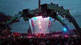 A concert stage; four large legs curve up above the stage and hold a video screen which is extended down to the band. The legs were lit up in green. The video screen has multi-coloured lights flashing on it. The audience surrounds the stage on all sides.