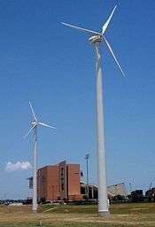 A photograph of two wind turbines in front of an empty college football stadium