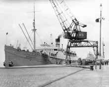 A photograph of a ship tied up to a pier. The bow of the ship points toward the left of the frame. On the right of the frame is an overhead crane.