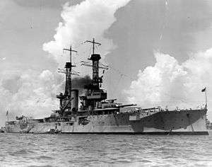 A large light gray battleship sits in harbor, dark black smoke billows lazily from the central smoke stacks