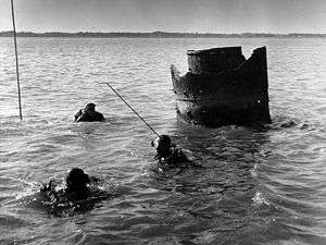 Remains of metal cylinder, 4-6 feet in diameter, protrude from water; three divers in foreground