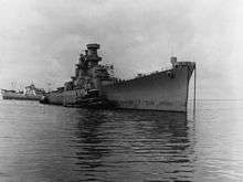 A clearly unfinished warship, with an incomplete superstructure and no gun turrets, is being moved by tugboats.