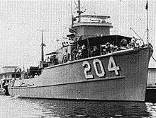 A black-and-white photograph of the USS Thrush floating near other structures. The boat is facing the camera with its prow facing the right of the photo, displaying "204" painted on its starboard hull.