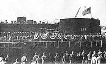 A large number of people are gathered around a platform erected just in front of a submarine.